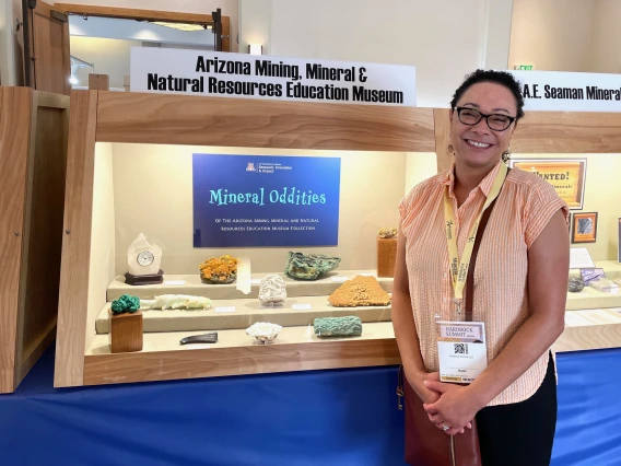 Director Marta Bones in front of our case of minerals at the 2024 Denver Gem and Mineral Show.